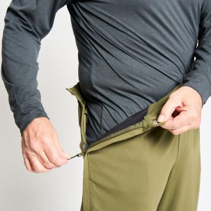 The torso of a man in a long sleeved, gray shirt, demonstrating the side zipper of a pair of khaki pants.